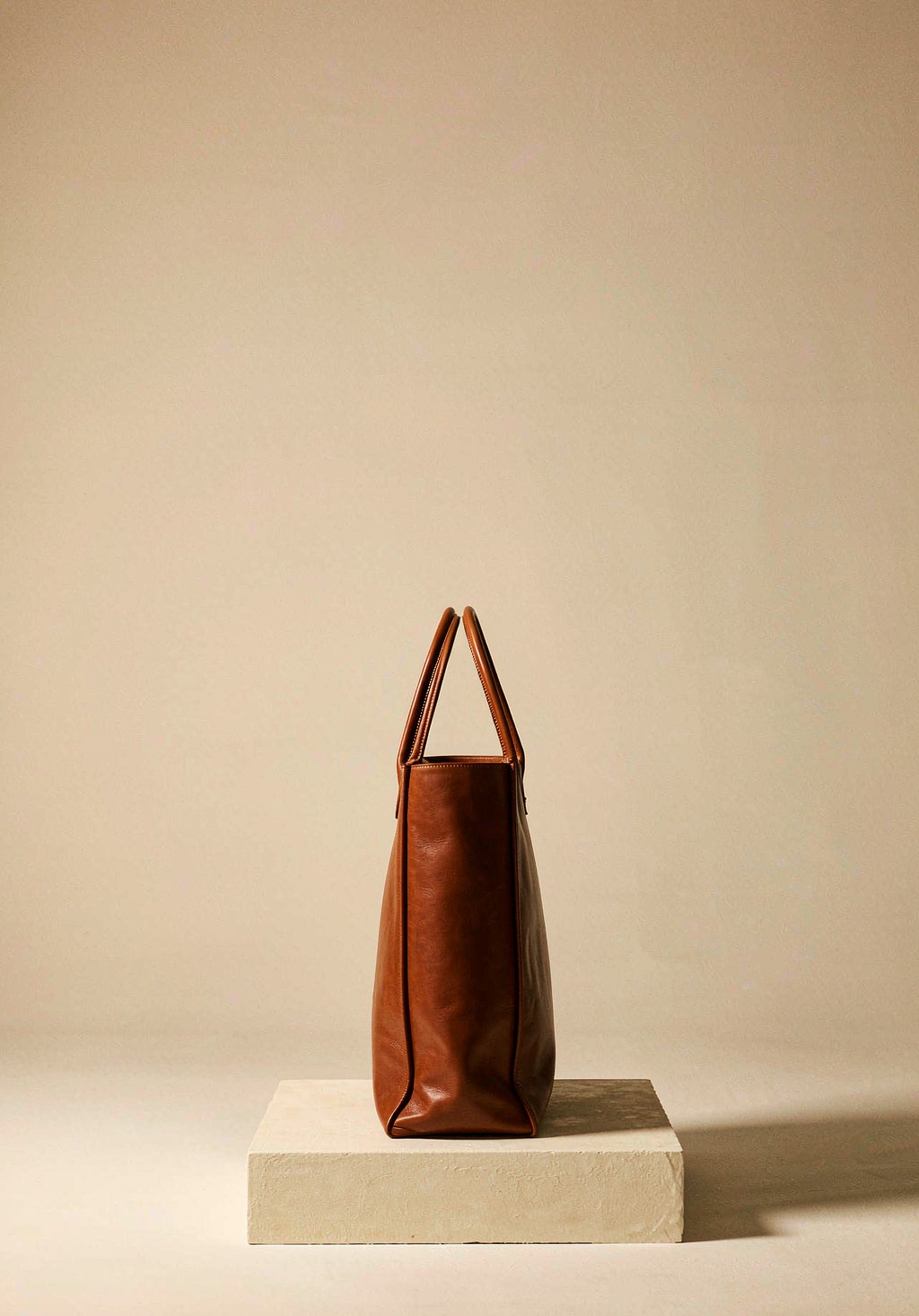 A tall structured cognac leather tote bag standing on a cream plaster plinth